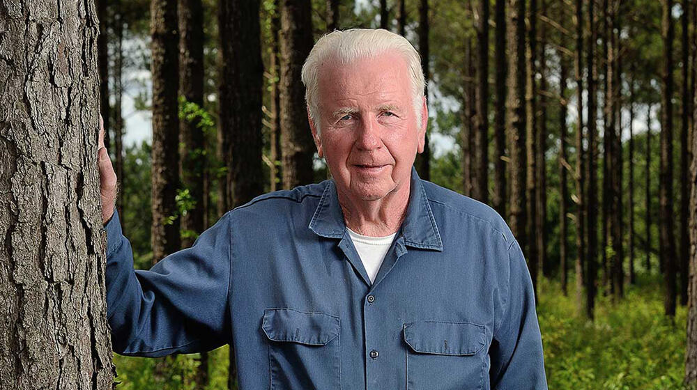 A white-haired male wearing a denim shirt and jeans stands beside a tree and smiles.