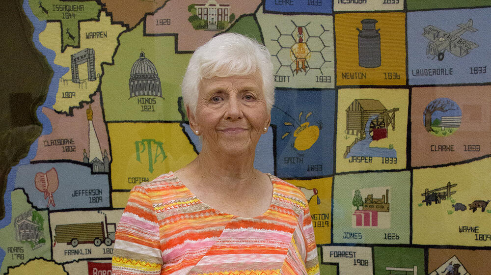 An elderly woman wearing an orange striped shirt stands in front of a large, multicolored, needlepoint county map of Mississippi.