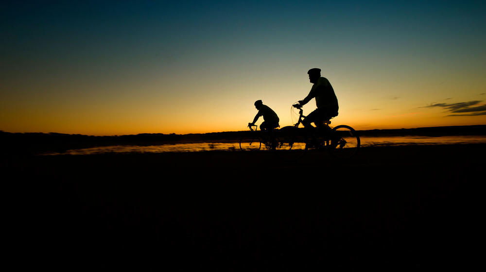 The silhouettes of two cyclists are seen in front of a blue and yellow sunrise that reflects on a lake below.