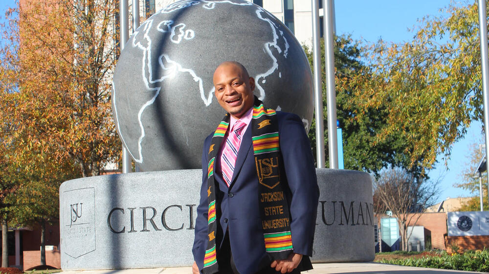 A man wearing a Jackson State University scarf and a navy suit jacket smiles in front of a sculpture on the JSU campus.
