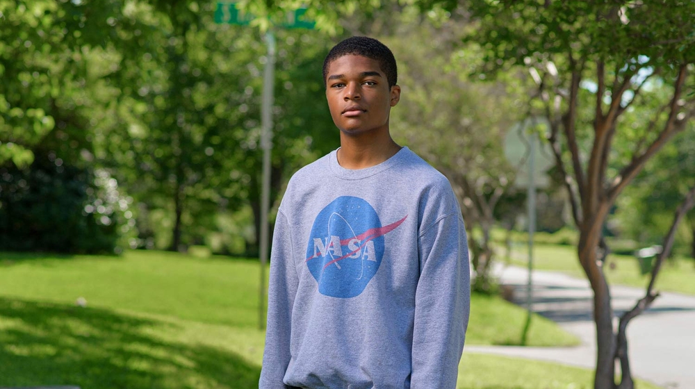 A young boy wearing a NASA sweatshirt stands on a sidewalk holding a camera by his side.