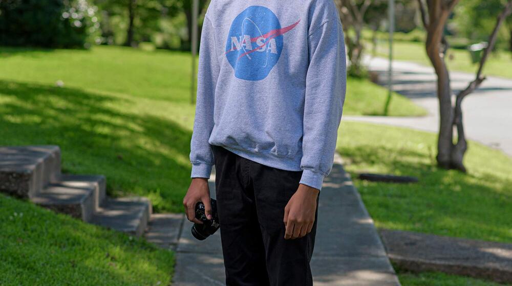 A young boy wearing a NASA sweatshirt stands on a sidewalk holding a camera by his side.
