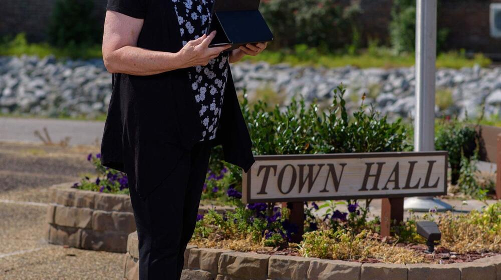 A smiling woman holding a tablet stands outside in front of a flagpole.