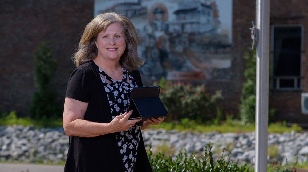 A smiling woman holding a tablet stands outside in front of a flagpole.