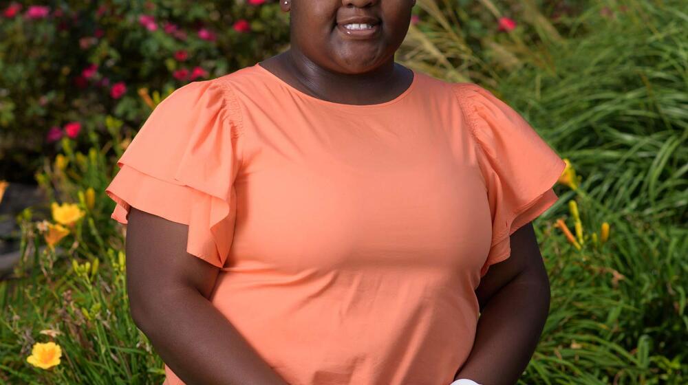 A smiling young girl wearing an orange shirt sits on a ledge in front of flowers.