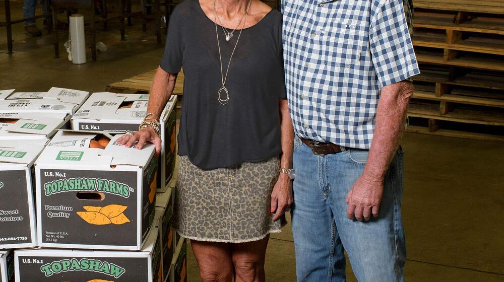 A man and woman stand next to a crate stacked with boxes of sweet potatoes.