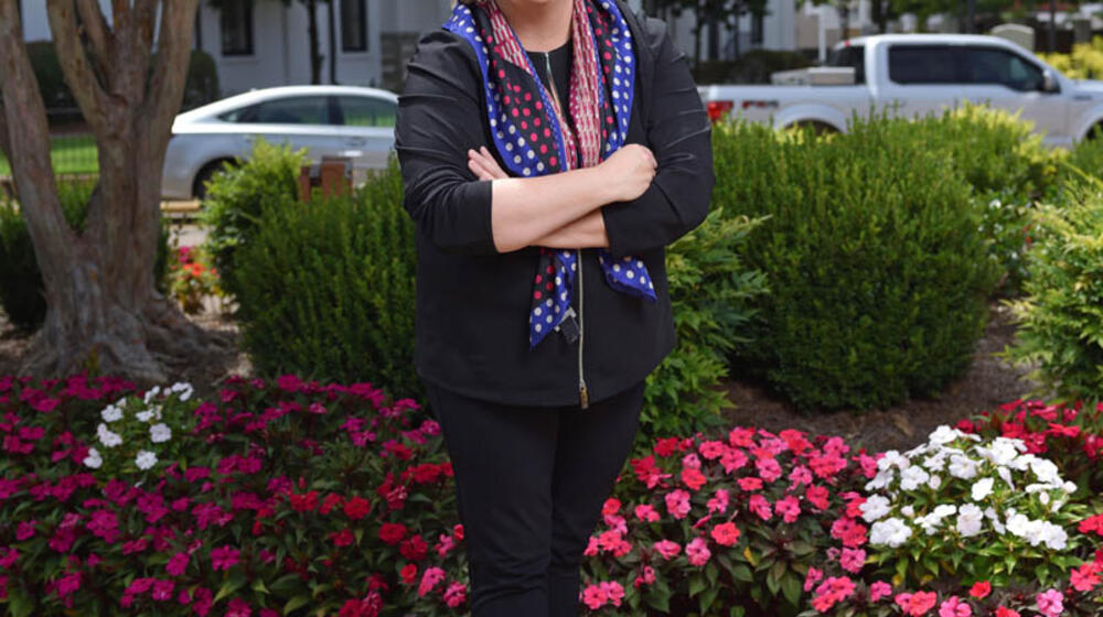 A woman, wearing all black clothes and a red, white, and blue scarf, smiling in front of flower bushes with her arms crossed.