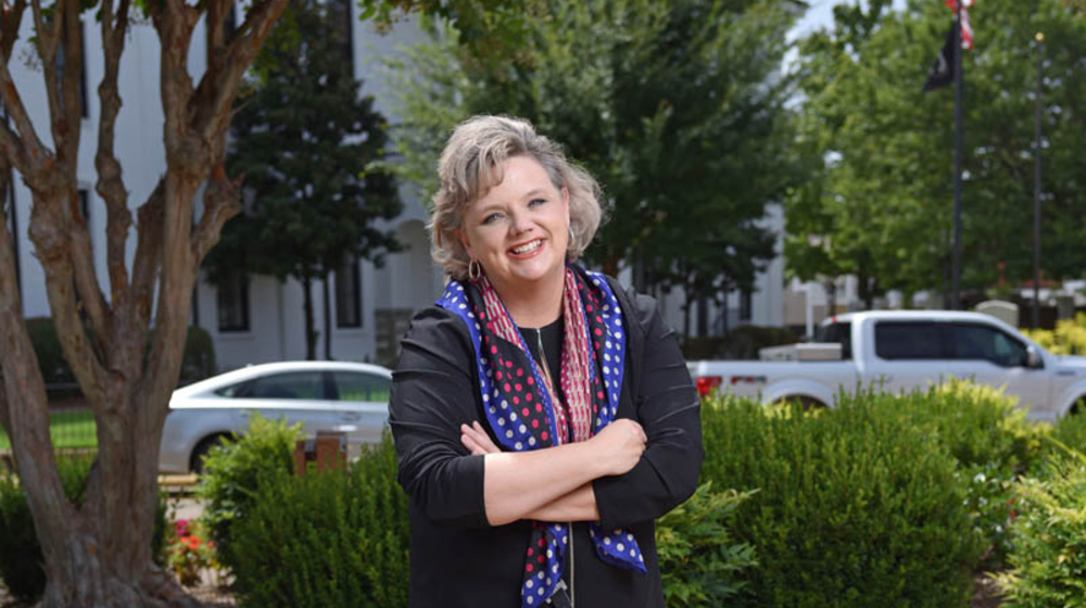 A woman, wearing all black clothes and a red, white, and blue scarf, smiling in front of flower bushes with her arms crossed.