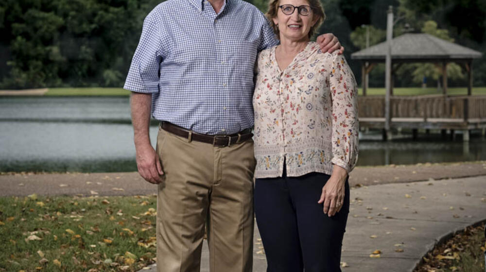 A man with his arm around a smiling woman standing on a sidewalk in front of a lake.