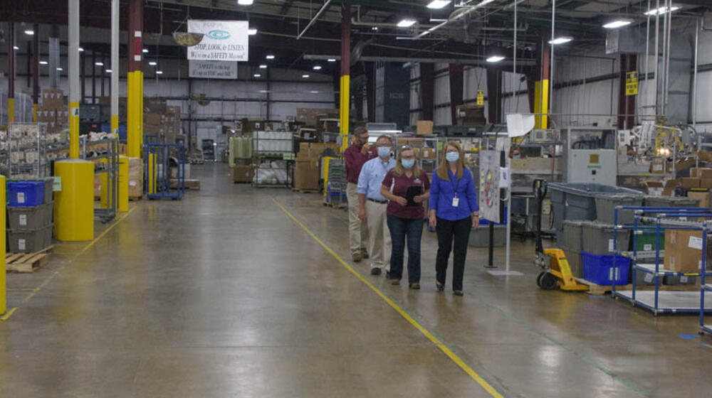 A line of four people walking in a room full of machinery and boxes.