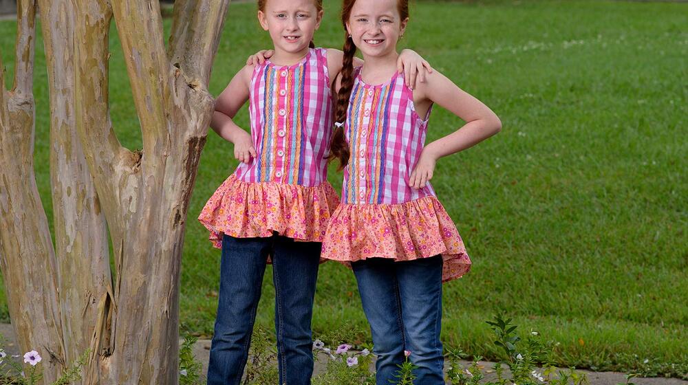 Two young girls with red hair and matching pink shirts stand next to each other with a hand on the hip.
