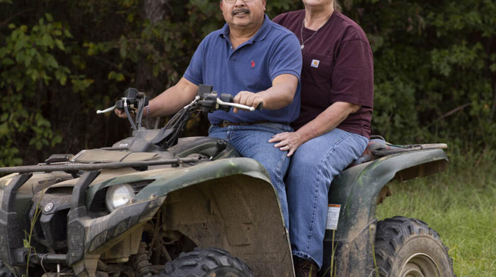 A man wearing a blue shirt and a woman wearing a maroon shirt sitting on a four-wheeler in tall green grass in front of dark green trees.