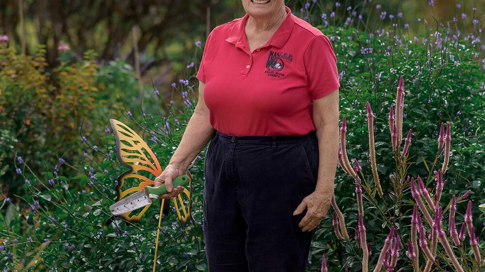 A woman wearing a red collared shirt standing in tall green grass and flowers. She holds a shovel in her right hand, which rests in front of a metal butterfly garden decoration.