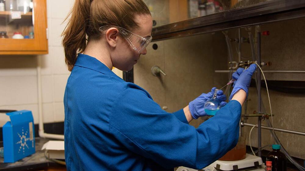 A young woman wearing safety goggles and a blue lab coat working in a lab.