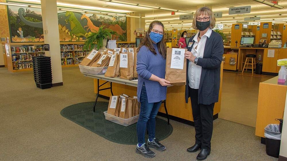 Two masked women with glasses and brown hair holding a brown bag.
