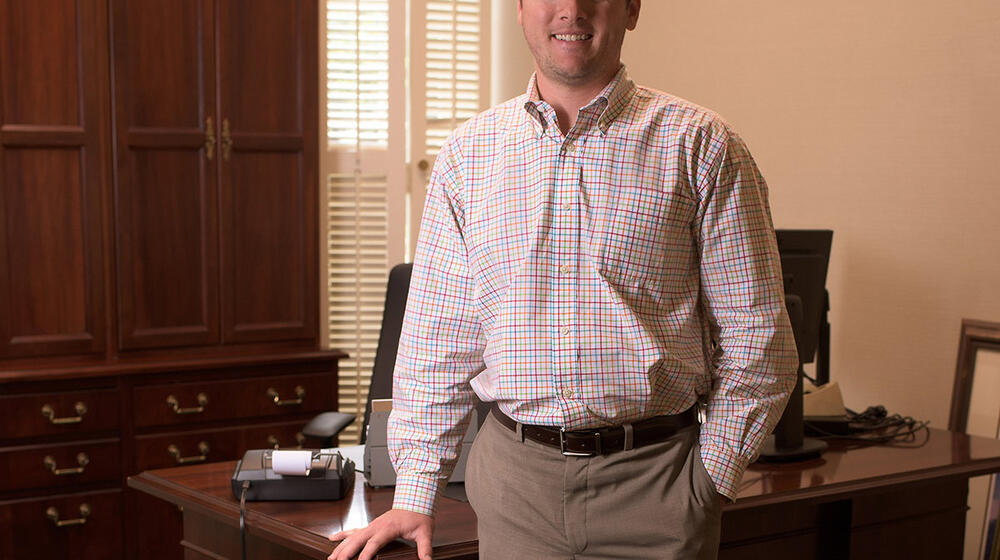 A man wearing a colorful plaid shirt stands in front of a desk.