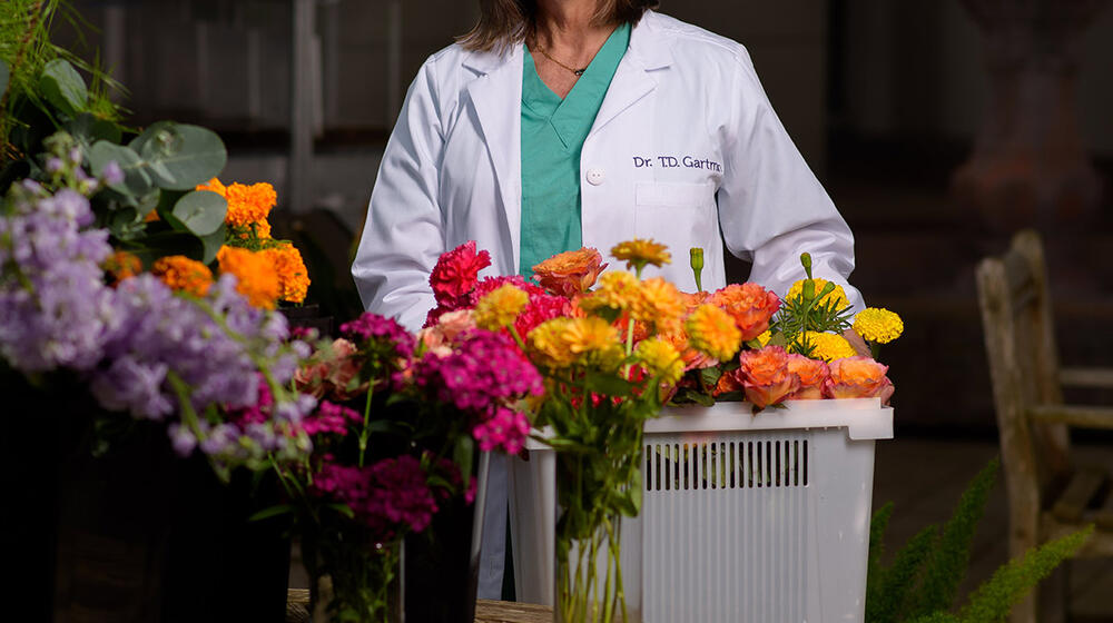 A woman wearing a white coat stands behind floral arrangements and a bucket full of flowers.