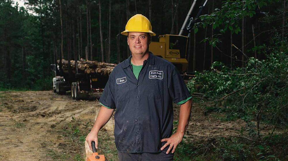 A man wearing a yellow hard hat and holding a chain saw stands in front of a trailer holding harvested timber.