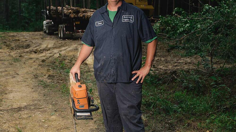 A man wearing a yellow hard hat and holding a chain saw stands in front of a trailer holding harvested timber.