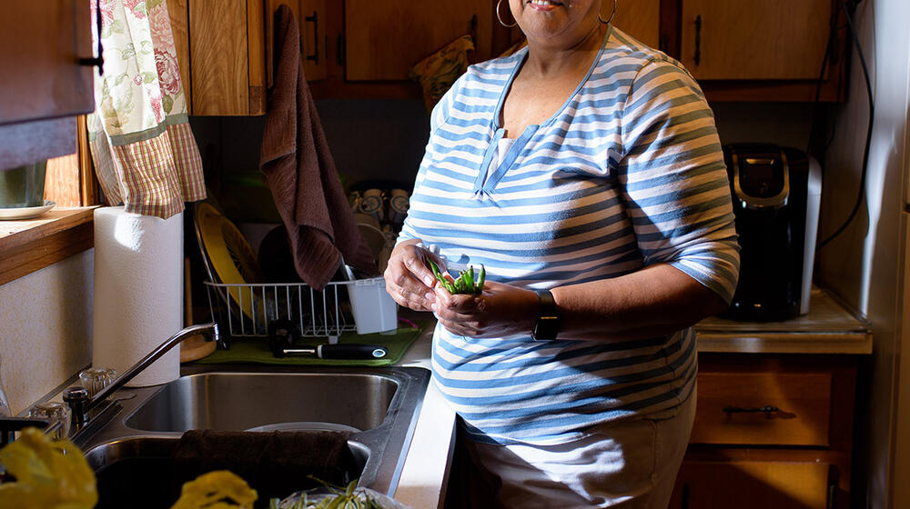 A woman wearing a white and blue striped shirt and holding fresh vegetables stands in front of a kitchen sink.