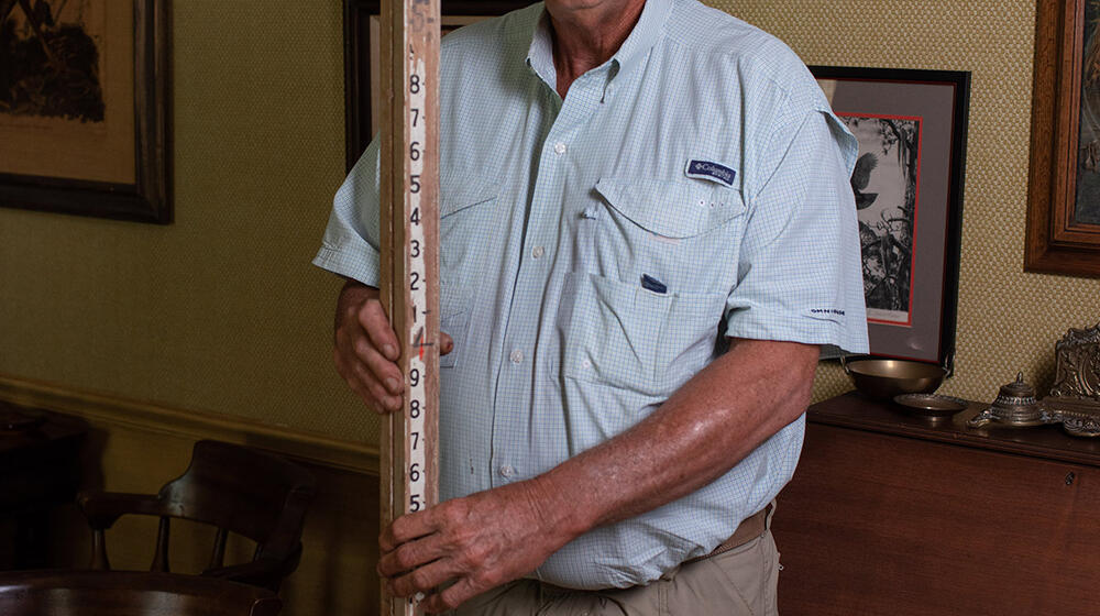 A man holding a large wooden measuring stick stands in front of a wall with several framed photos.