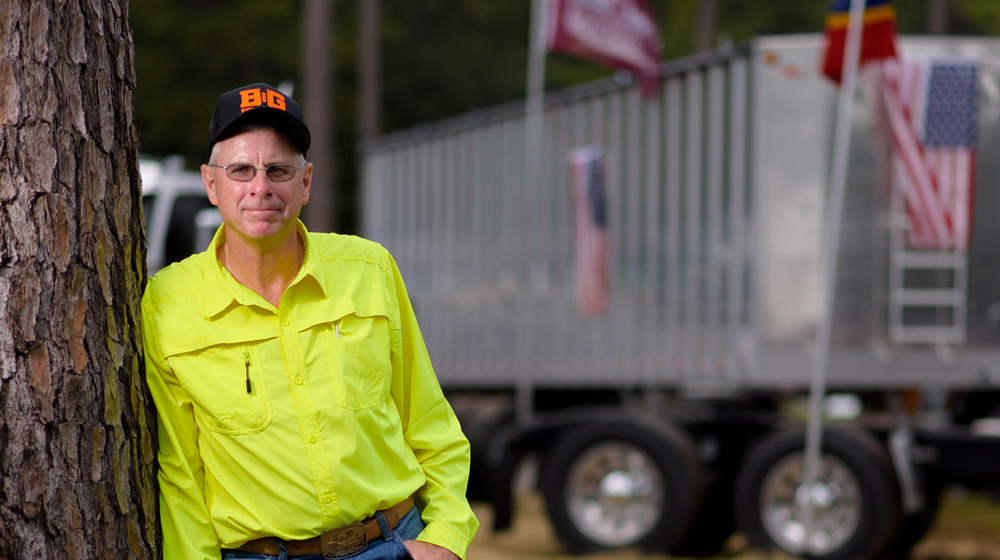 A man wearing a bright yellow button-up shirt leans against a large truck and rests one hand in his blue jean pocket.