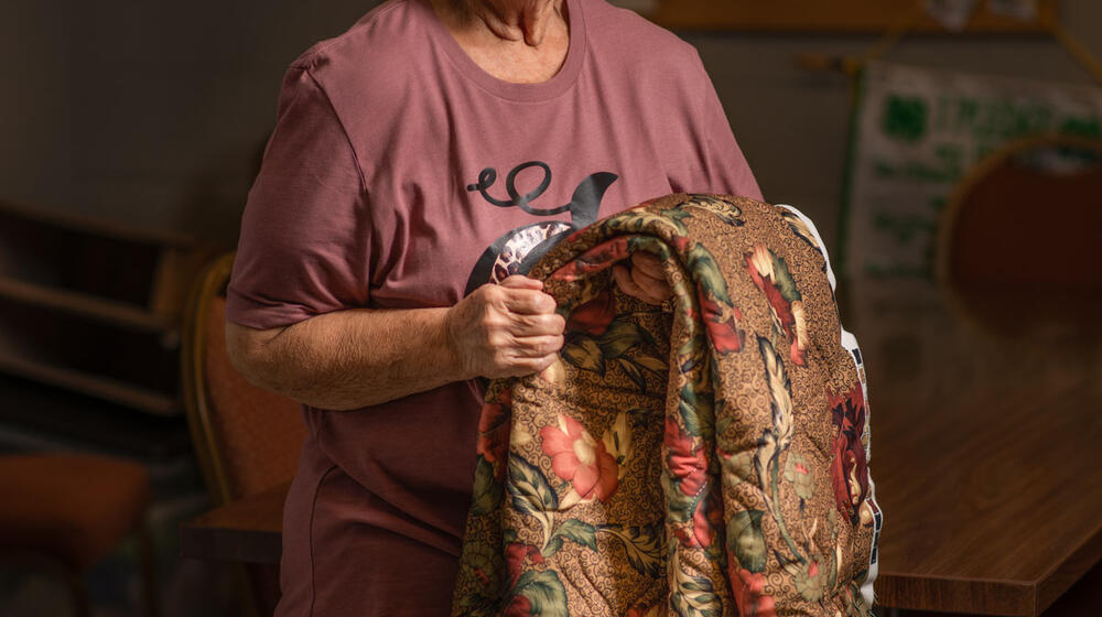 An older woman smiles while holding a quilt.