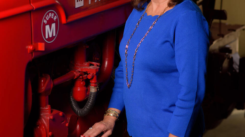 A smiling woman wearing a blue shirt stands next to and rests her arm on a red piece of machinery. 