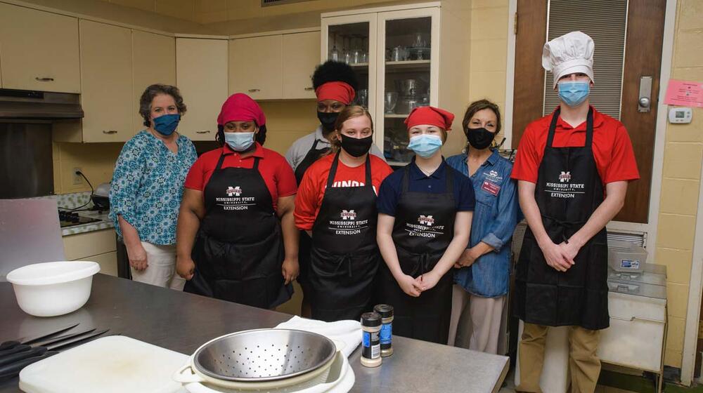 Five young adults and two women wearing black aprons and face masks smile for a group photo in a kitchen.