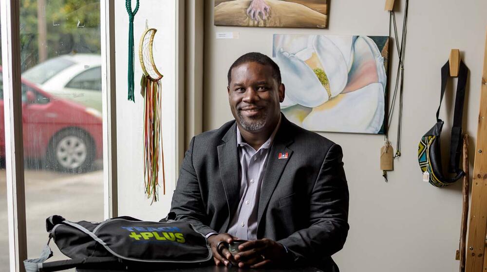 A man wearing a suit sits at a round table with a small mug and a bag that says “Teach Plus.”
