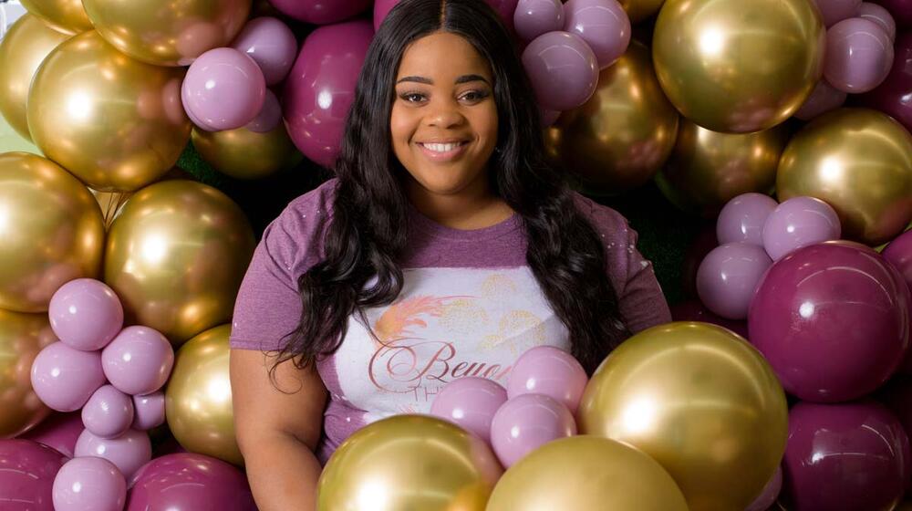 A smiling young woman wearing a shirt that reads “Beyond the Arch” stands next to several balloons and jars of party supplies.