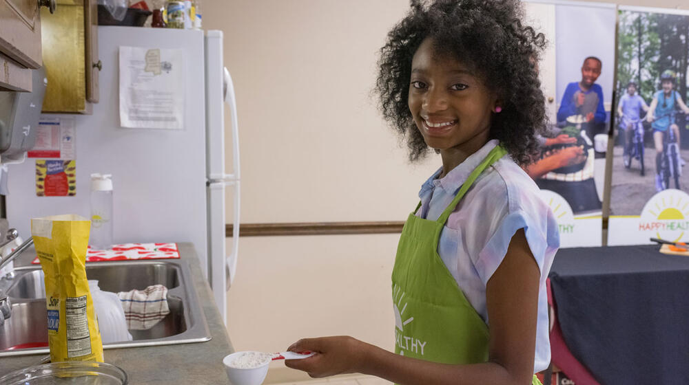 A smiling young girl wearing a green apron holding a measuring cup full of flour.