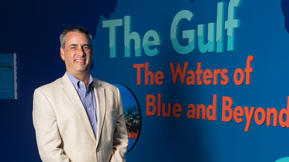 A man wearing dress clothes stands in front of a blue wall and smiles with his hands clasped in front of his body.