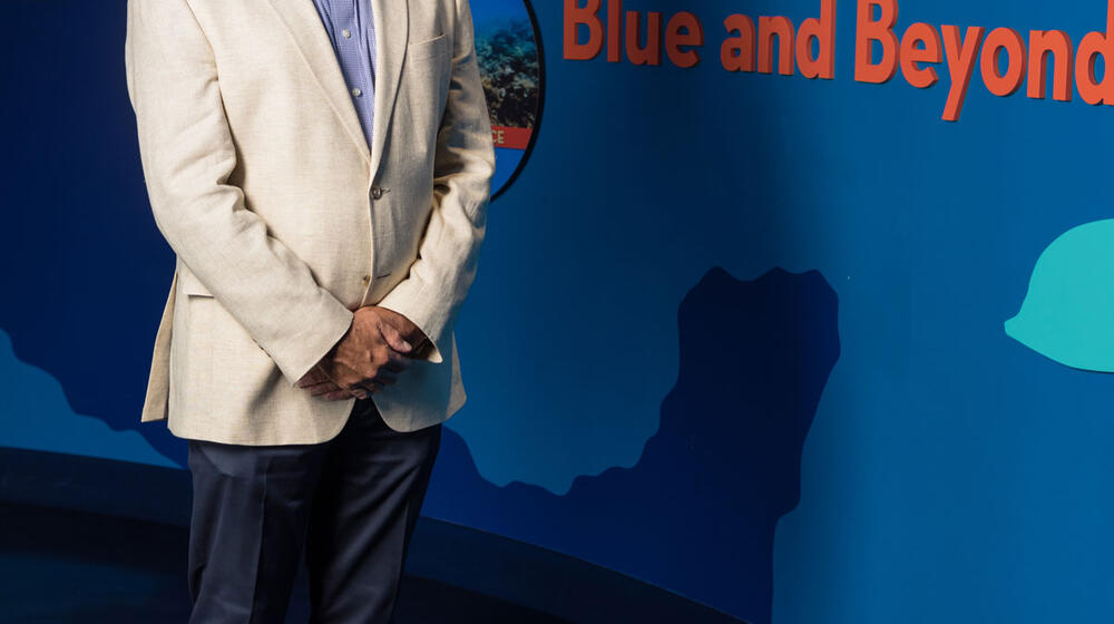 A man wearing dress clothes stands in front of a blue wall and smiles with his hands clasped in front of his body.