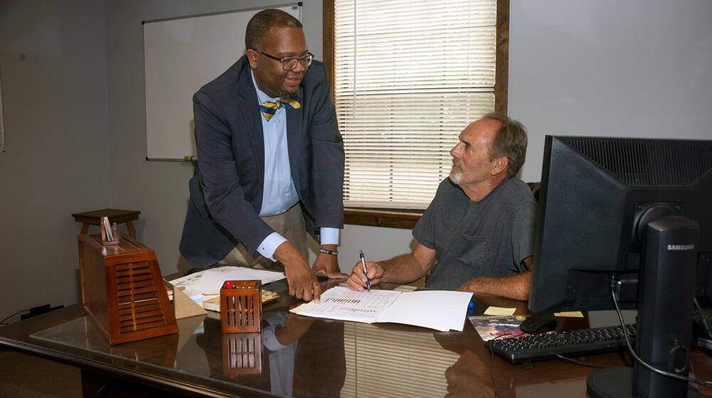A standing Black man points to a paper on a desk that a seated white man with a pen also points to.