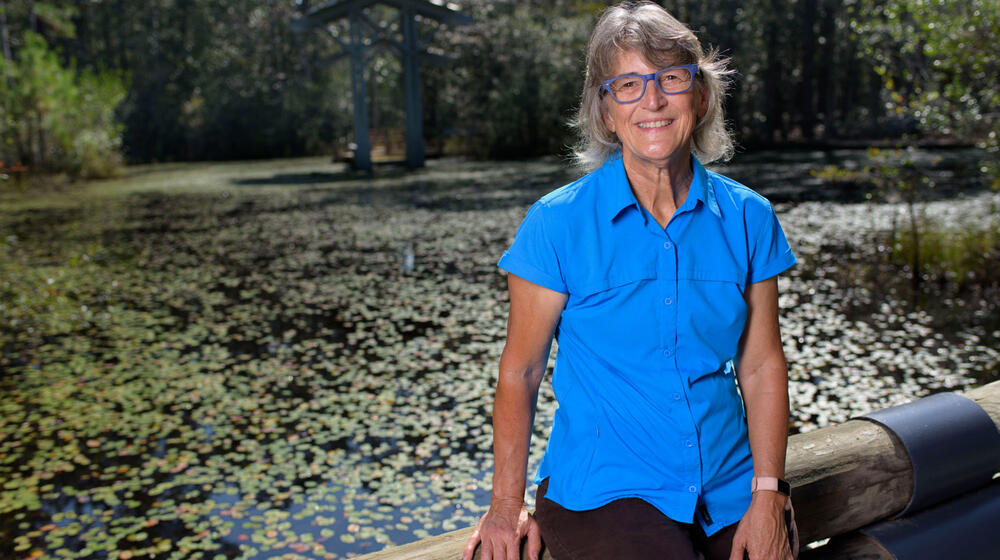 A woman, smiling, wearing a blue polo and sitting on a wooden fence.