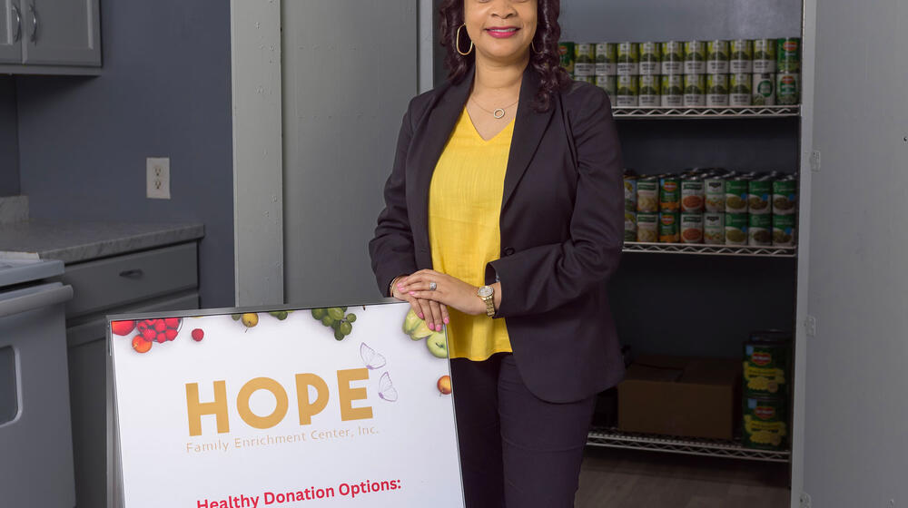 A professionally dressed woman standing in front of many rows of food cans and beside a sign that lists “HOPE.”