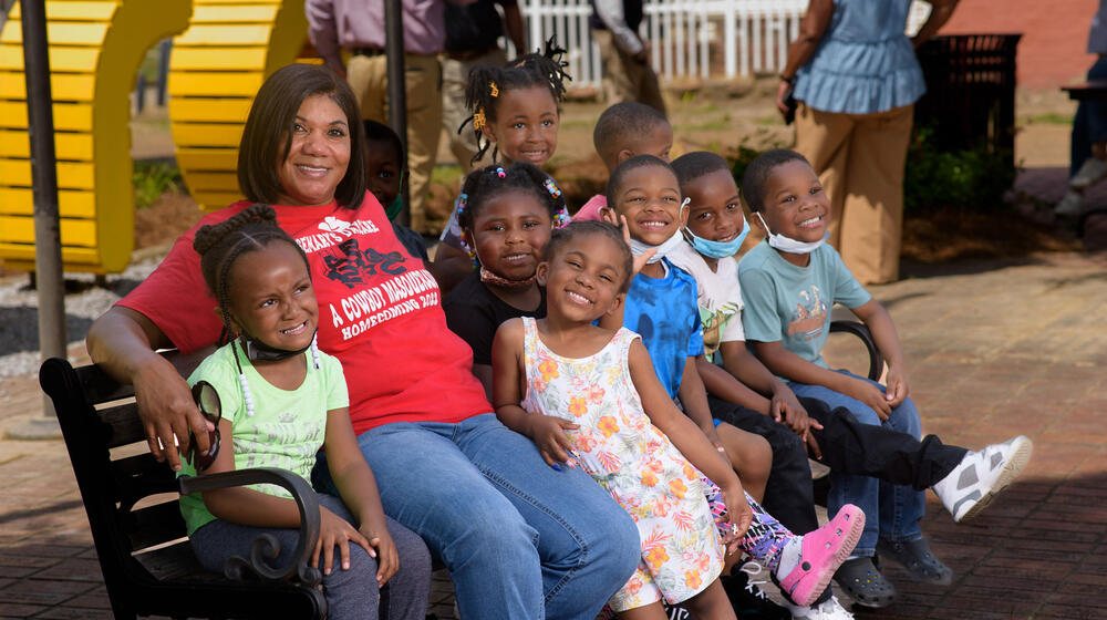 A woman, wearing a red T-shirt listing Rosemary’s Daycare on it, smiles while sitting on a bench with many small children sitting beside her, also smiling.