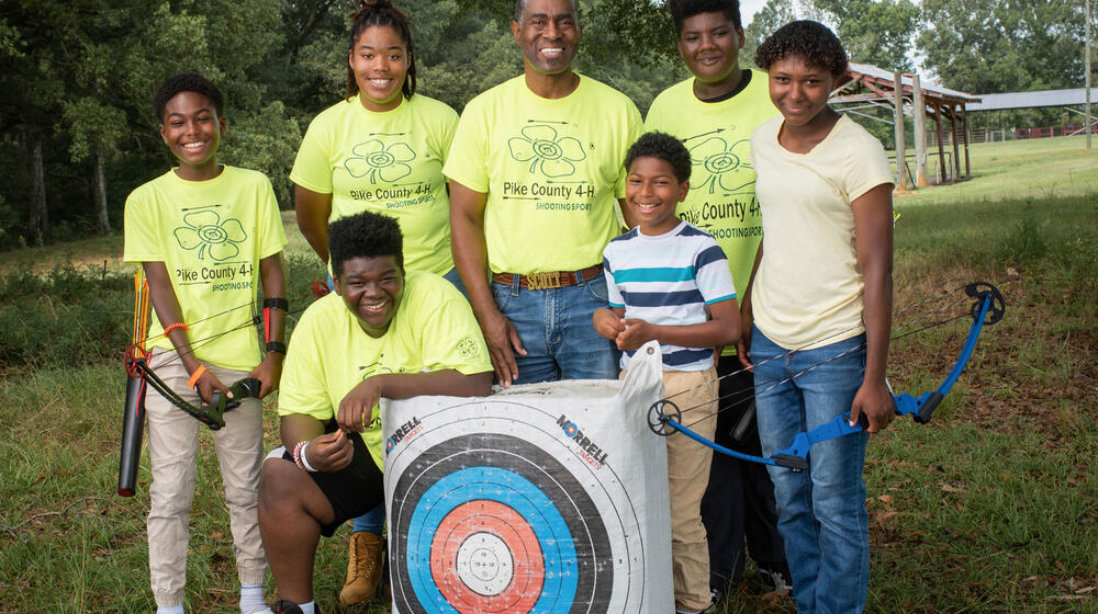 A man standing behind an archery target and surrounded by six children, all smiling.