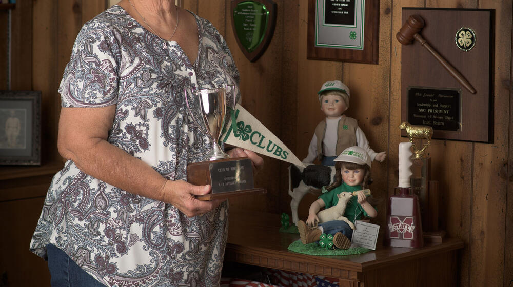 A woman, smiling, holding a 4-H flag and 4-H trophy and standing in front of several 4-H plaques affixed to the wall.