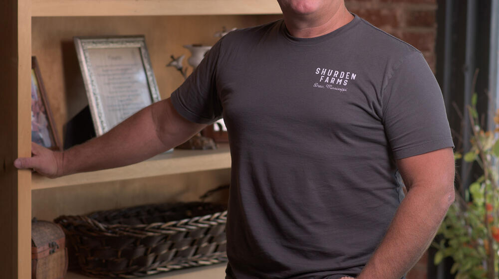 A blonde man wearing a T-shirt with Shuden Farms listed on it standing in front of a bookshelf, smiling.