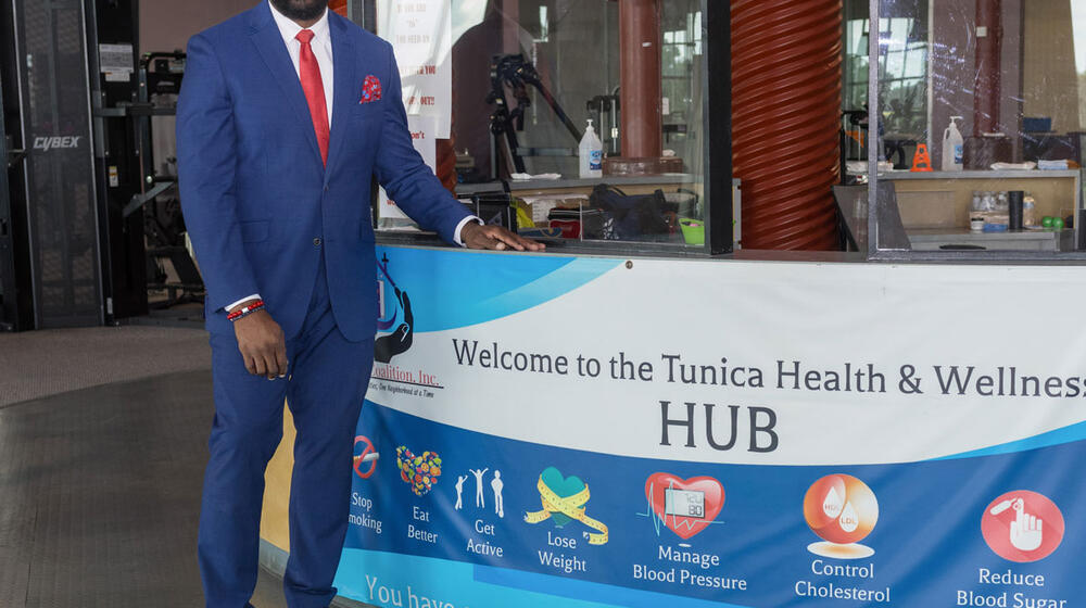 A man wearing a suit stands beside a desk with a banner listing “Welcome to the Tunica Health & Wellness Hub.”