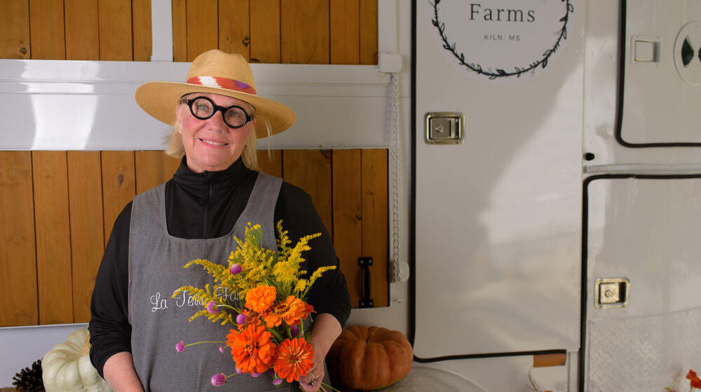 A woman with a straw hat and round-framed glasses holding a bunch of flowers and smiling.