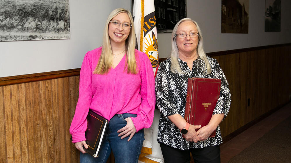 Two women standing in front of a flag, smiling.