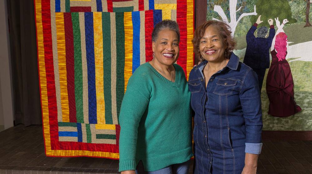 Two Black women smiling and standing in front of a hanging multicolored quilt.