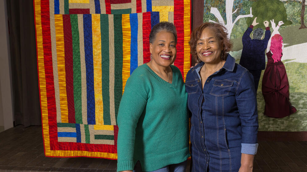 Two Black women smiling and standing in front of a hanging multicolored quilt.