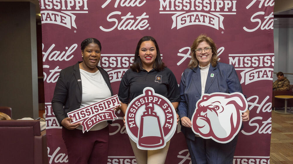 Three women standing in front of a maroon and white Mississippi State University banner.