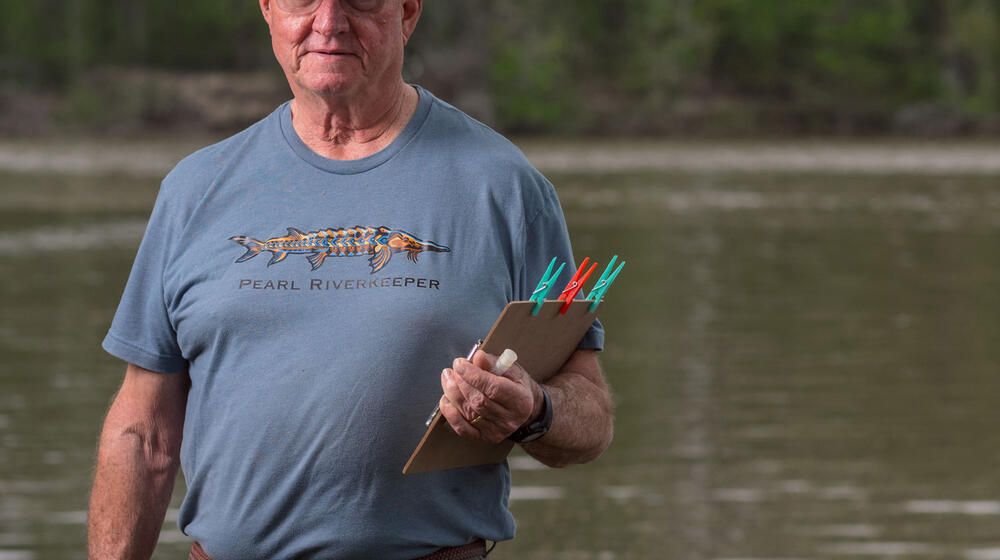 A man with a blue visor and T-shirt listing “Pearl Riverkeeper” and holding a clipboard in front of a waterway.