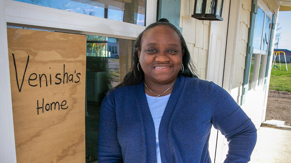 A woman standing beside the window of a house that has a sign with “Venisha’s Home” listed on it.