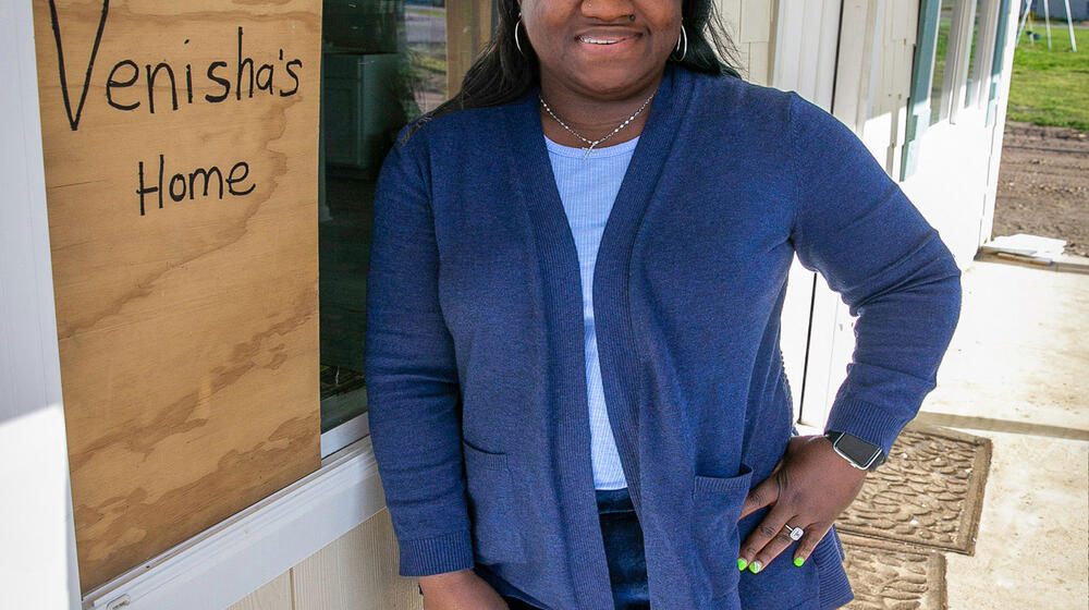 A woman standing beside the window of a house that has a sign with “Venisha’s Home” listed on it.